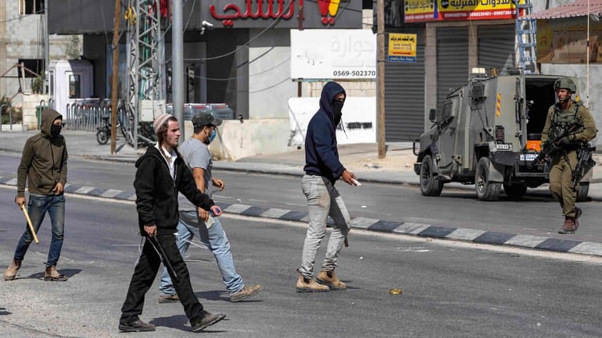 Colonos sionistas encapuchados, armados con palos y piedras, cruzan una calle de Ḥuwwāra, en Cisjordania, mientras un soldado israelí observa junto a un vehículo militar