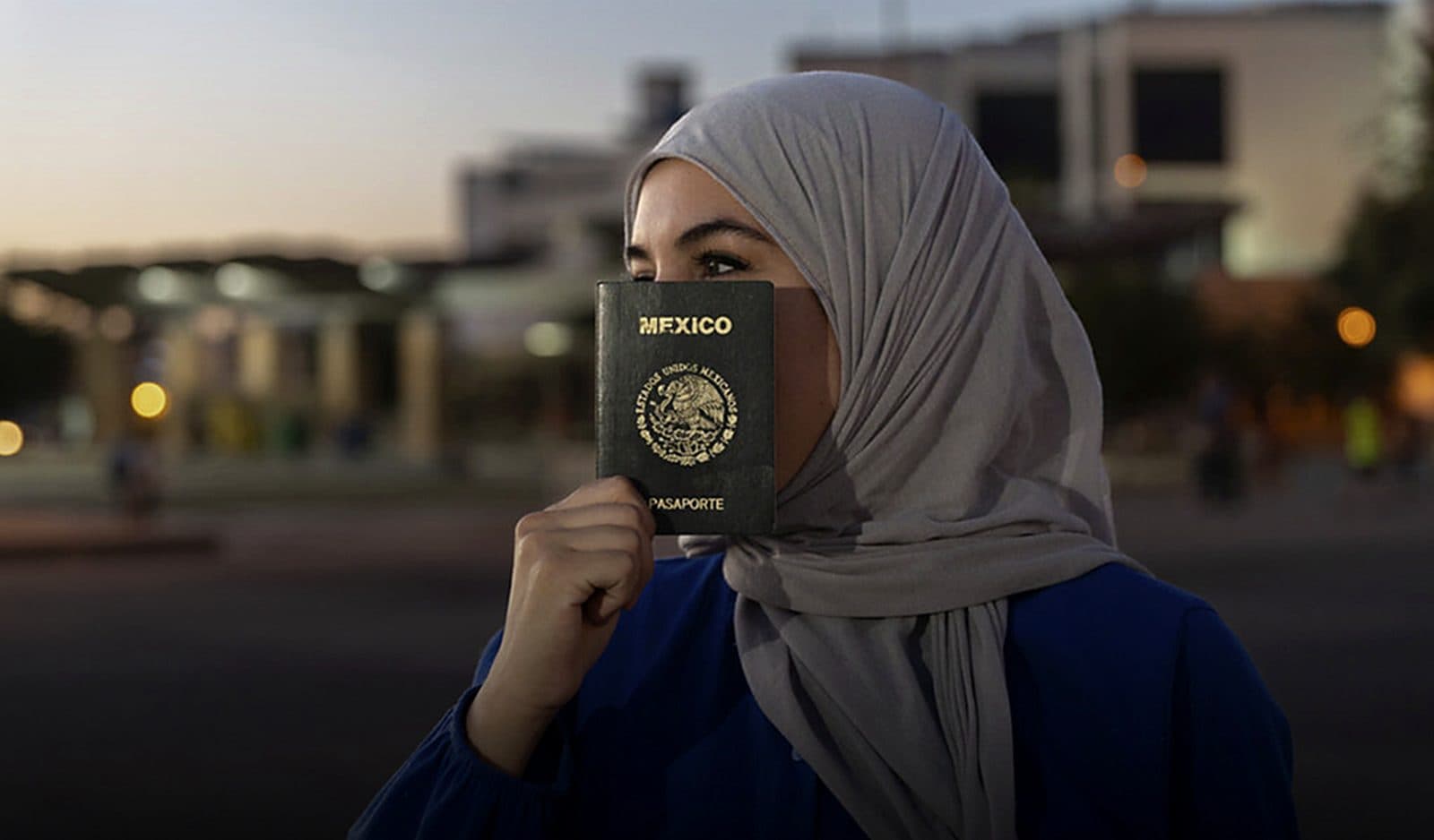 Mujer con hiyab gris y blusa azul sostiene un pasaporte mexicano frente a la parte baja de su rostro, en una calle al atardecer con edificios y luces desenfocadas al fondo